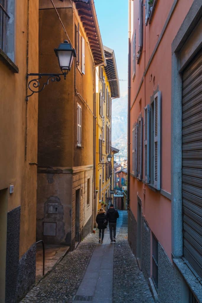 Two people walking side by side in one of Bellagio's alley. All buildings are pastel colored, mainly terracotta and orange.