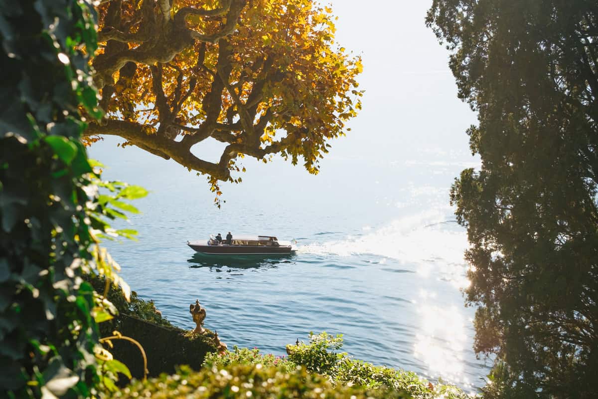 A wooden boat passing in front of Villa Balbianello, framed by trees with yellow and orange leaves during autumm