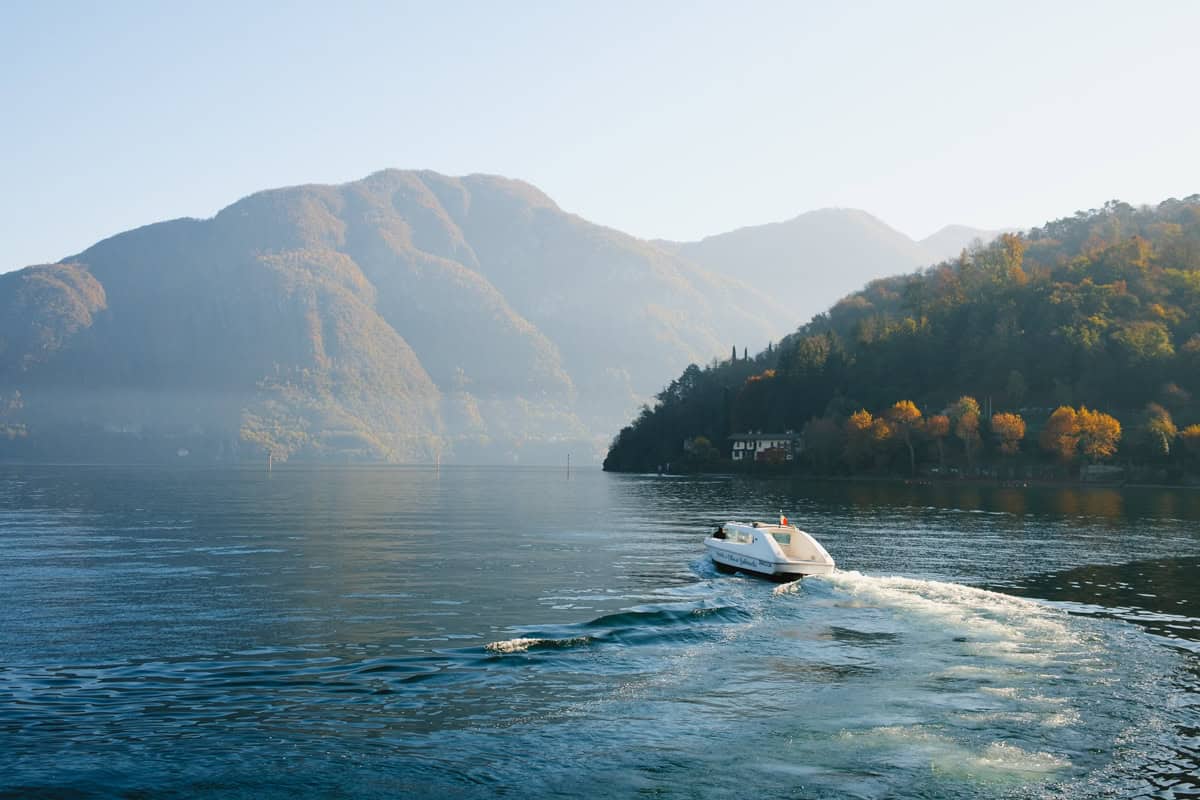 A small white taxi boat headed towards villa Valbianello, navigating during a misty morning in a hazy light, with foliage colors in the hills around the lake