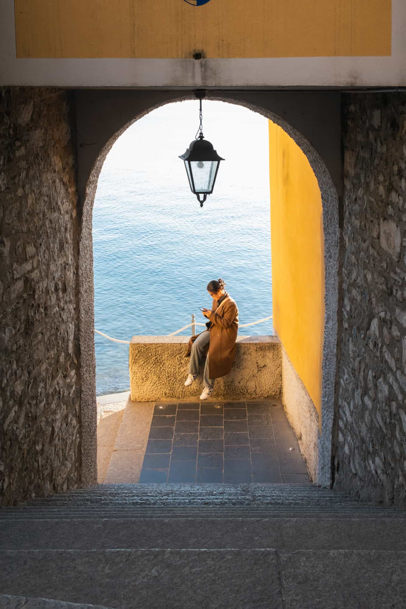 A woman in a long coat looking at her phone and sitting on a ledge under an archway overlooking Lake Como in Varenna, with sunlight illuminating the yellow wall behind her.