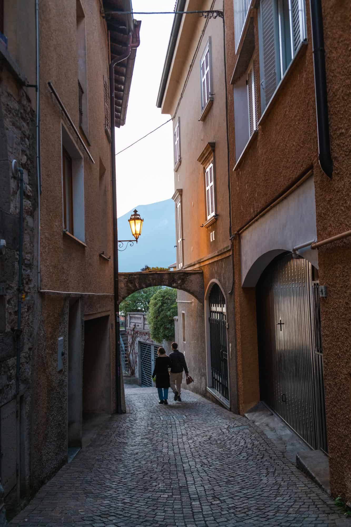 A narrow cobblestone alley in Varenna, framed by rustic buildings and a glowing streetlamp, with a couple walking toward the end of the street