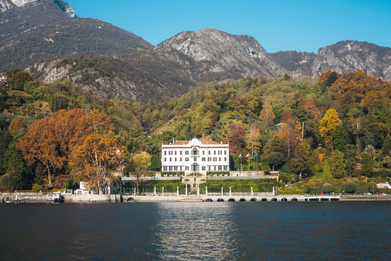 The white building of Villa Carlotta in Tremezzo surrounded by trees and foliage colors, photographed from the lake