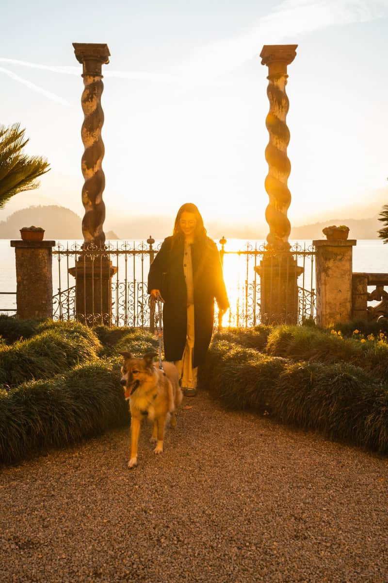 Gaia walking her dog along a gravel path in Villa Monastero at sunset, framed by intricate stone pillars and glowing golden light behind her