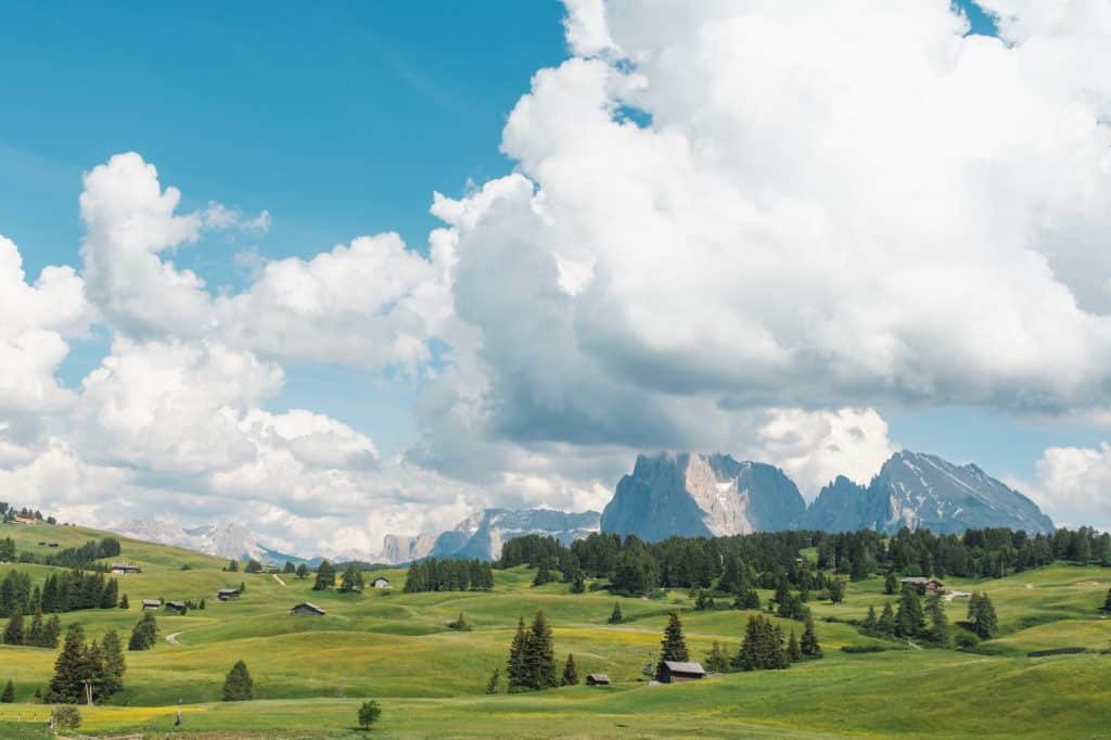 A view of alpe di siusi, with wooden chalets scattered among the fields. There are big white clouds in the sky partially covering the mountains in the back.