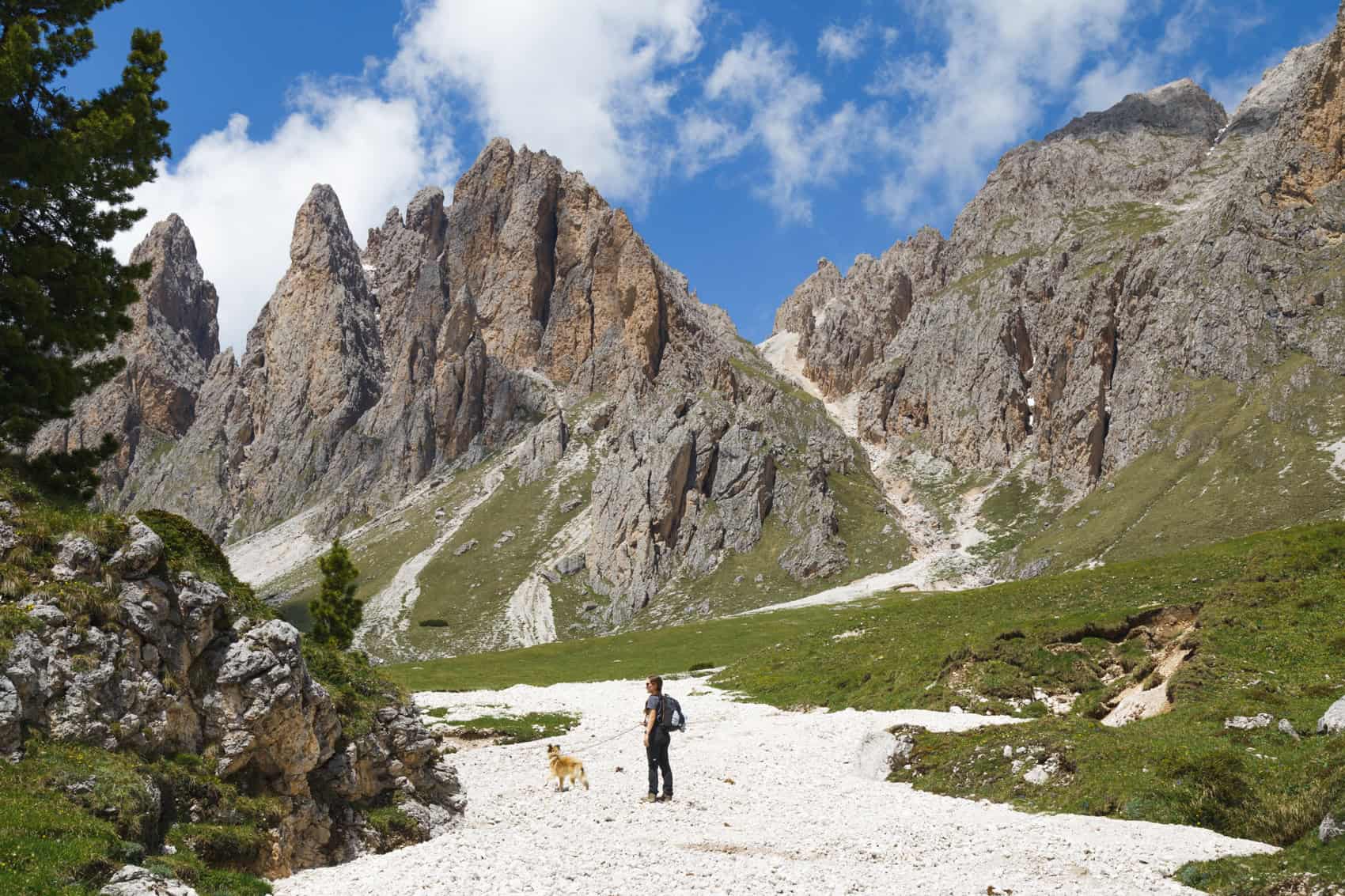 A woman walking on a white trail in the dolomites with her dog and big mountains in the back