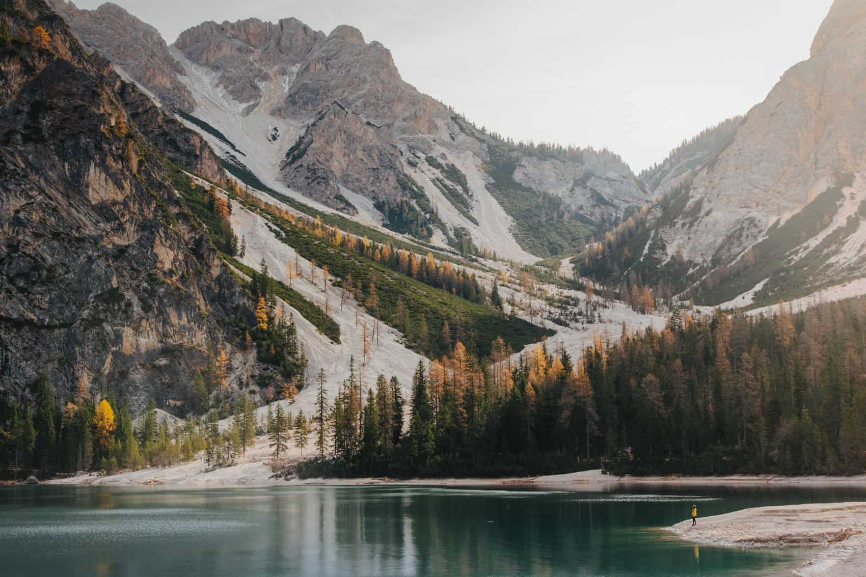 A landscape photo of Lake Braies, with mountains reflecting on the lake and a very tiny Gaia in the corner wearing a yellow coat.