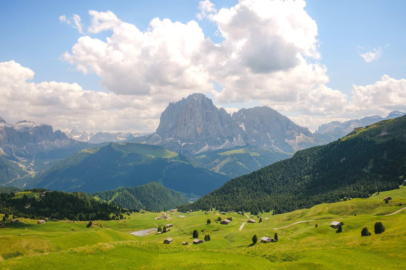 Sassolungo shot from Seceda on a summer day with a blu sky, big white clouds and green pastures with little wooden houses in the front of the picture