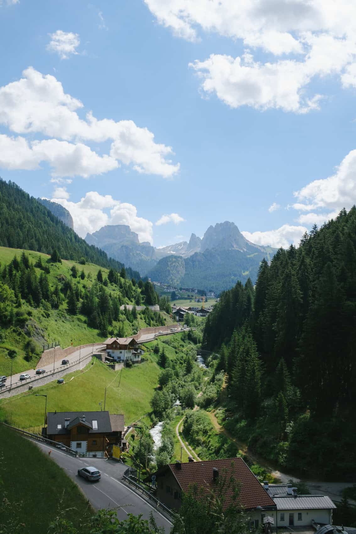 Cars passing on a road in Val Gardena with mountain peaks in the background and a forest on the right side