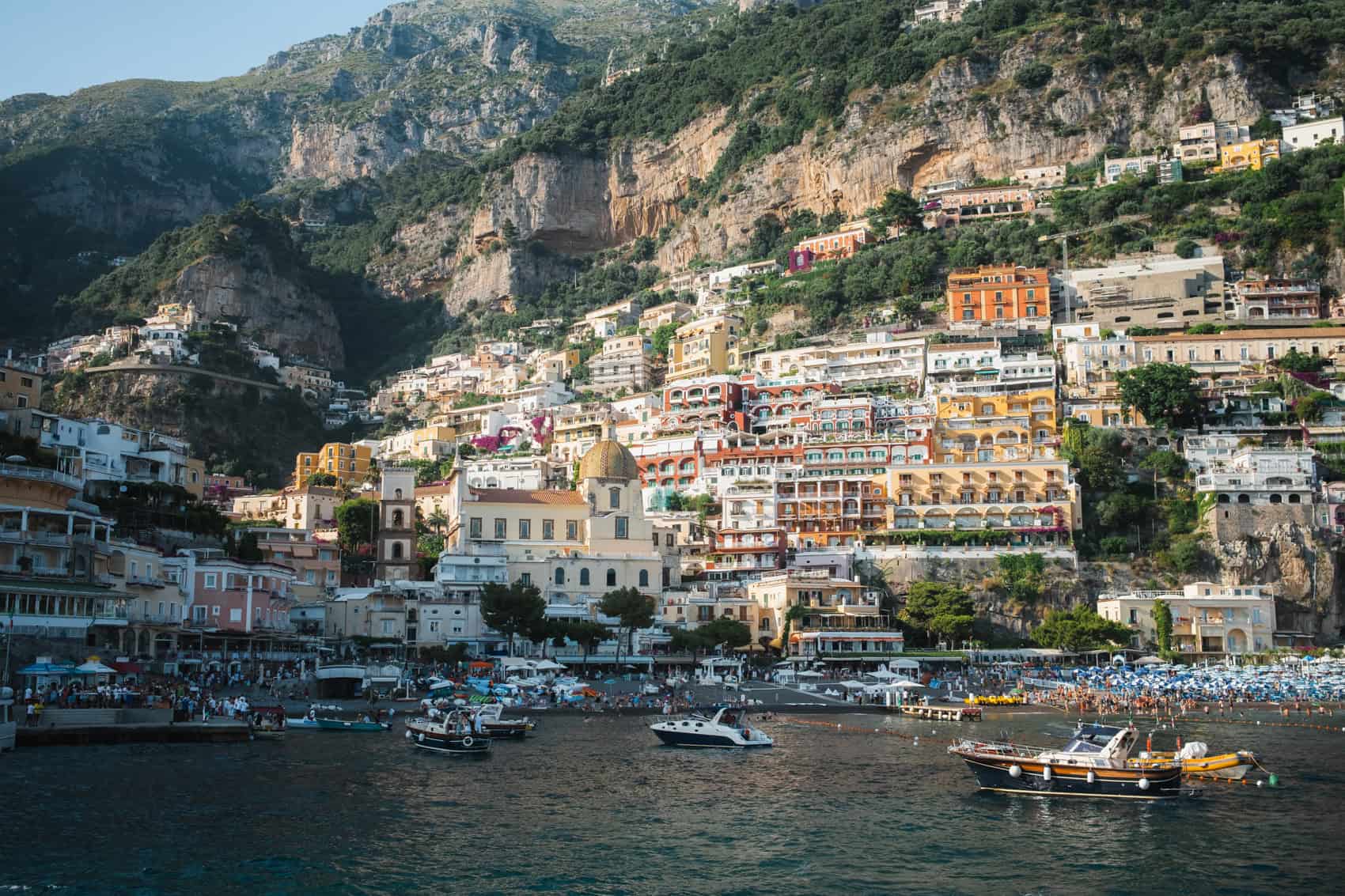 Positano in the Amalfi coast is one of the best places to visit in italy for first-timers, here is seen from the water, during a boat tour. There are several small boats in front of the town, which is illuminated in gold light a bit before sunset