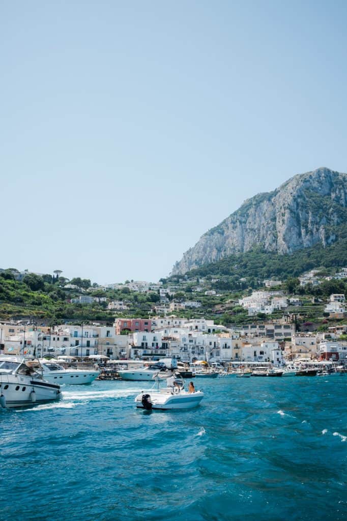 Capri seen from the water during a boat tour. The water is crystal clear, an unreal bright blue, and there are boats in front of the seafront, with plenty of small white buildings