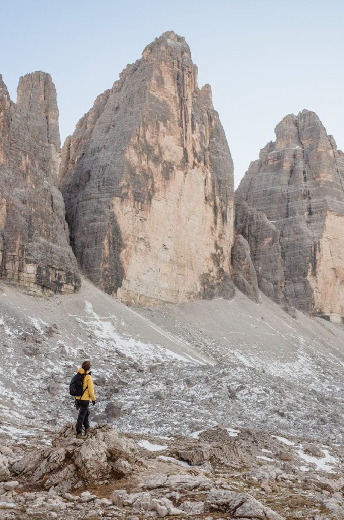 Gaia hiking in a yellow jacket standing on rocky terrain, gazing up at Tre Cime di Lavaredo in autumn.