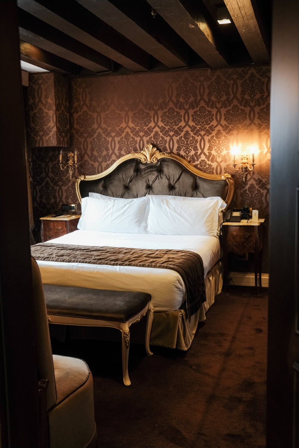 A room at Hotel Nani Mocenigo Palace. The walls are covered in brown elegant fabric and the bed is following the traditional venetian decor with a thick golden frame.