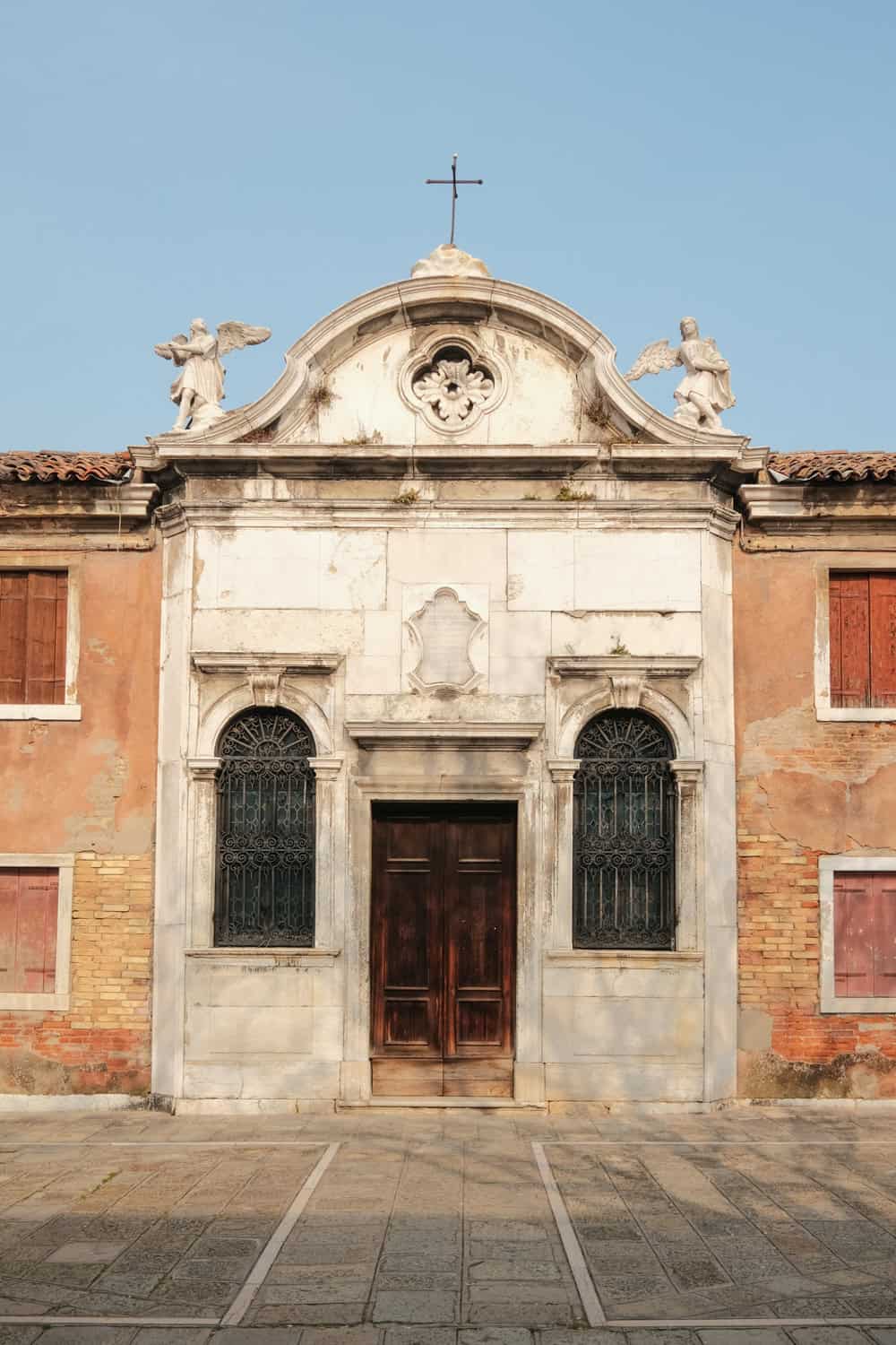 Front view of a weathered historic church facade in Murano, featuring a marble entrance with two arched windows, a wooden door, and angel statues flanking a small cross on the roof. The surrounding terracotta walls and shuttered windows highlight the aged, rustic charm of the island’s architecture.
