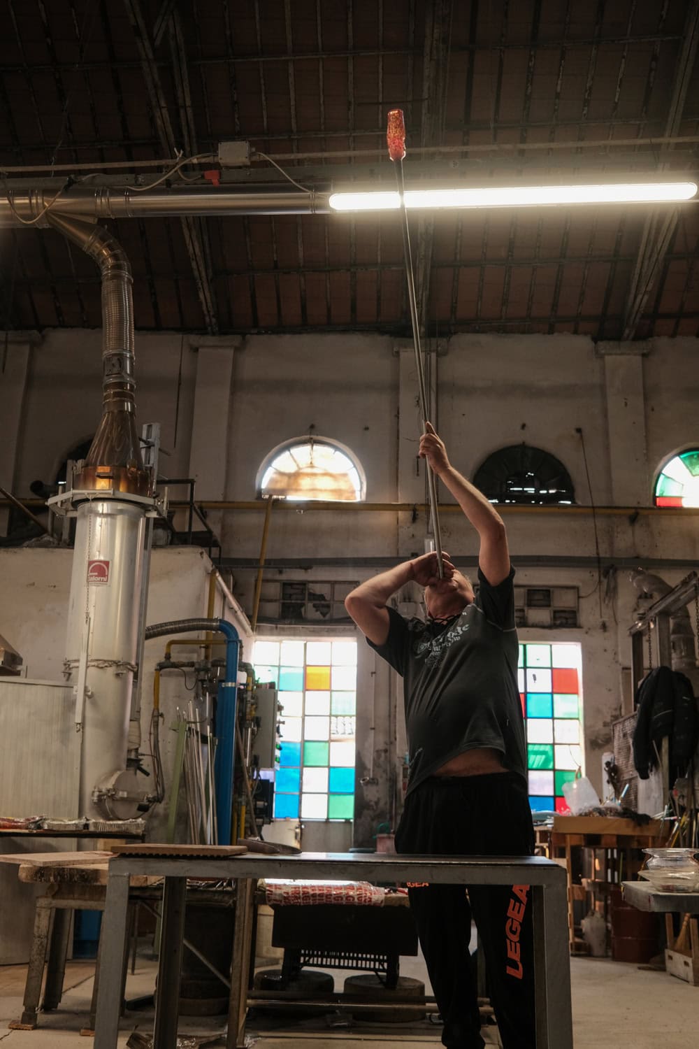 A glassblower in Murano shapes molten glass at the end of a long pipe inside a traditional workshop. The dimly lit studio features industrial equipment and colorful stained-glass windows that add a pop of light behind the artisan.