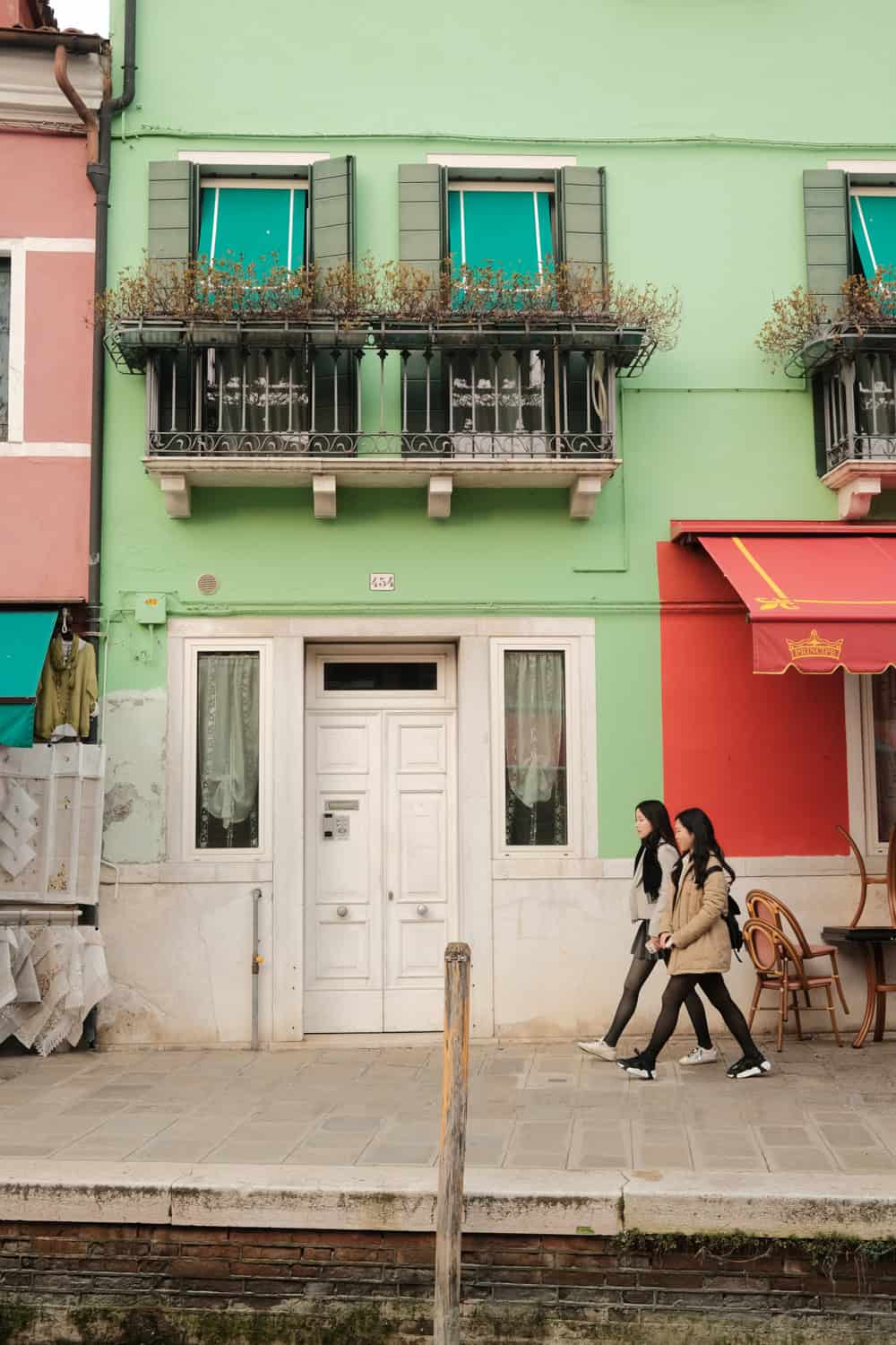 Two women walk past a row of colorful houses in Burano, Italy, with a mint-green building front and bright red awning. The scene is framed by shuttered windows, delicate balcony railings, and canal-side pavement, capturing the island’s signature bold palette and relaxed pace.