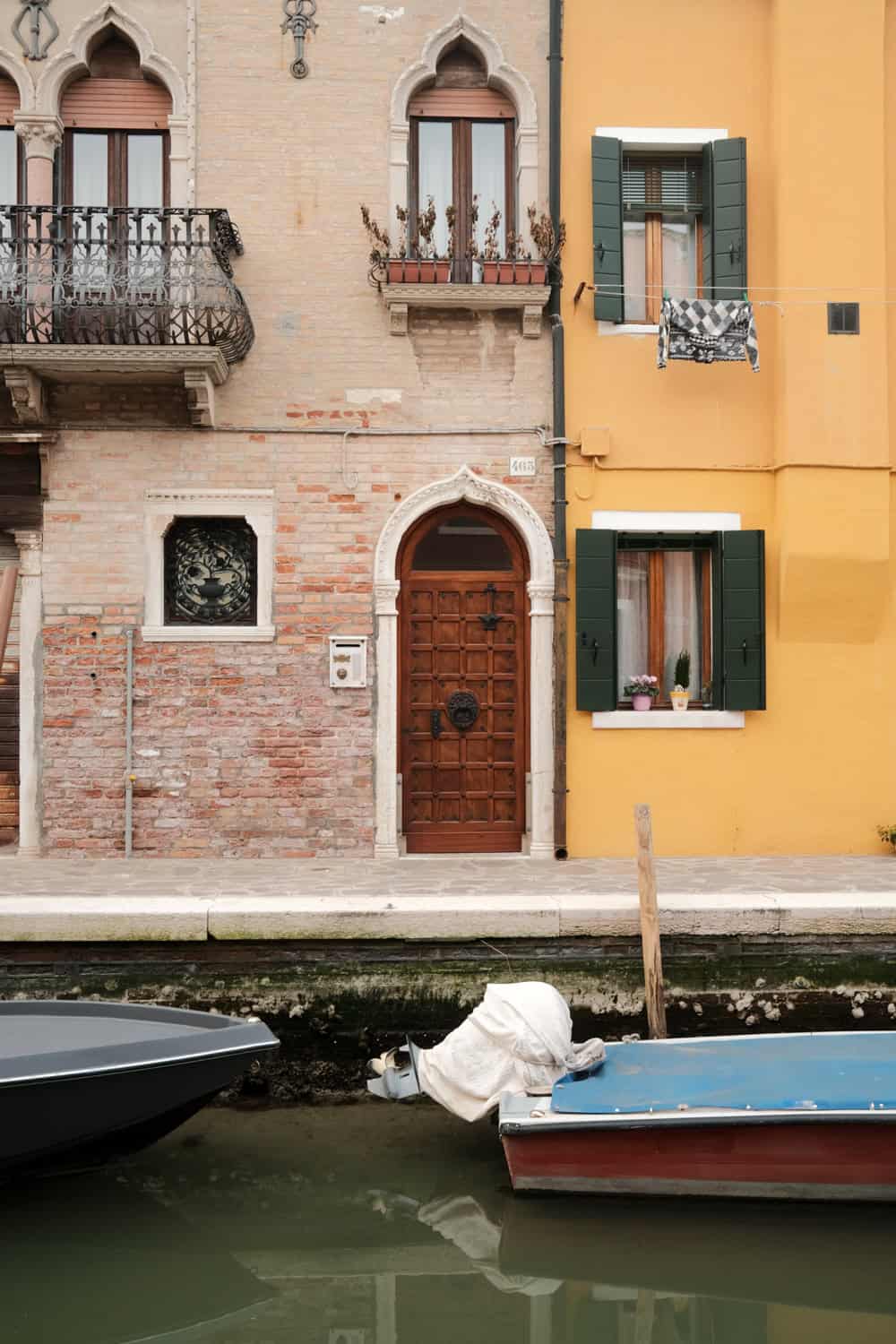Charming canal-side buildings in Burano, Italy, featuring a mix of exposed brick and golden yellow facades with arched windows, green shutters, and flower boxes. A pair of small boats are docked in the calm canal below, adding to the island’s picturesque and lived-in feel.