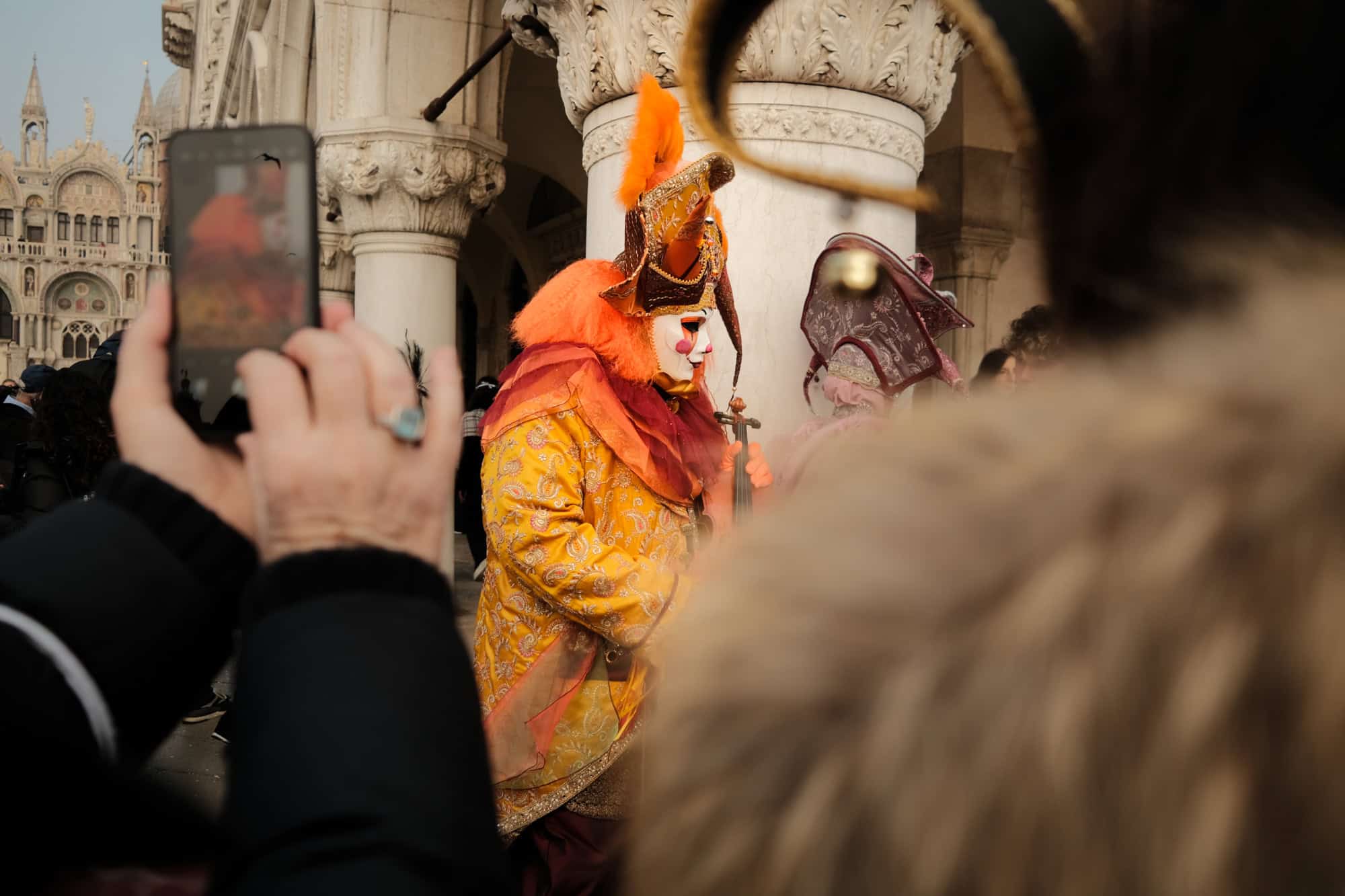 A man in an orange costume with a white mask walks among the crowd in st mark's square during venice carnival